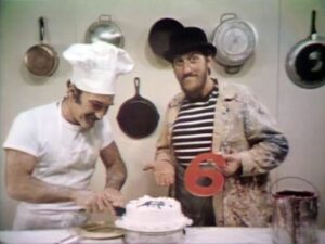 Still of the Mad Painter looking at a chef with a cake while holding a number 6